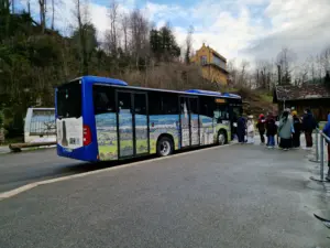 Blue open-top tour bus with passengers boarding at a stop near a wooded hillside building
